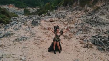 Karmaşık kostümlü çarpıcı bir figür kayalık arazinin ortasında duruyor. Karmaşık tasarımlar ve kalın renkler sergiliyor. Arka planda yeşilliği olan engebeli bir manzara var..