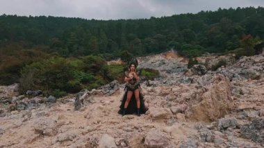 Çarpıcı bir figür, dalgalı bir pelerin ve dramatik bir kostüm giyen kayalık bir arazide güven içinde duruyor. Arka planda yemyeşil ağaçlar ve bulutlu bir gökyüzü var, mistik bir atmosfer yaratıyor..