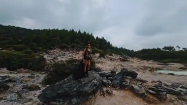 Karmaşık tasarımları olan dramatik siyah bir elbise giymiş bir figür, seyrek bitki örtüsü ve bulutlu gökyüzü ile çevrili kayalık bir arazide duruyor. Sahne bir gizem ve fantezi hissi uyandırıyor..