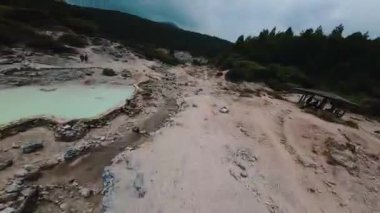 Bulutlu bir gökyüzünün altında, sık ormanlarla çevrili, kumlu bir yolu olan kayalık bir manzara. Arazi, doğal ve engebeli bir ortam yaratmak için çeşitli büyüklükteki taş ve yeşillik parçalarını barındırıyor..