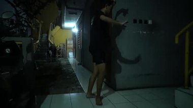 Gecenin karanlığında loş bir koridorda duran ve duvara gölge düşüren biri. Ayarda döşeme döşemeleri ve arka planda sarı bir duvar bulunan bir bina cephesi yer alıyor..