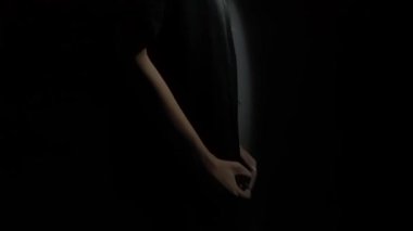 Karanlık bir ortamda duran, küçük bir ışık kaynağıyla aydınlatılmış bir insan. Bireyler dalgın görünür, başları hafifçe eğik ve elleri göğsünde, ışık ve gölge arasında dramatik bir kontrastlık yaratır..