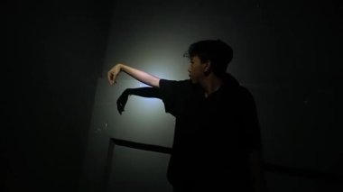 Loş ışıklı bir odada bacağını duvara dayayan bir insan, fenerle aydınlatılıyor. Sahne, minimalist bir ortamda odaklanma ve kararlılık hissi yakalar..