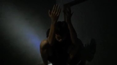 Karanlık bir odada oturan biri, elleri havada ve gölgeler dramatik bir etki yaratıyor. Atmosfer umutsuzluk ya da mücadele hissi verir..