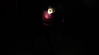 Bir el feneriyle aydınlatılmış, gölgeli bir figürün kol uzattığı loş bir sahne. Arka plan çoğunlukla karanlıktır, pencereden gelen belli belirsiz ışıkla..