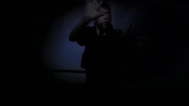 Karanlık bir ortamda genç bir çocuk, üzgün görünüyor ve iki eliyle başını tutuyor. Sahne, sönük bir ışıkla aydınlanır, dramatik ve gergin bir atmosfer yaratır..