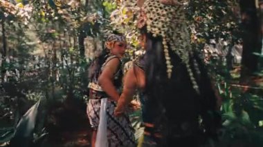 Arkadan iki kadının el ele tutuşmalarını takip eden zarif bir atış. Güzel, geleneksel Cava kostümleri giymişler. Manzaralı, doğal ve ağaçlık bir alanda ilerliyorlar..