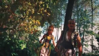 Geleneksel Endonezya kıyafeti giymiş neşeli bir adam ve kadın güneşli bir çam ormanında yürürken dans edip birlikte gülüyorlar. Enerjileri canlı ve mutlu. Aşkı ve kültürel mirası kutluyorlar..
