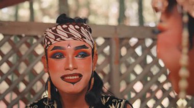 Tam teatral makyaj ve geleneksel batik başlık (blangkon) giymiş bir erkek Cava dansçısına odaklanan yakın çekim. Endonezya performans sanatını sergileyerek, etkileyici bakışlarla partneriyle iletişim kuruyor..