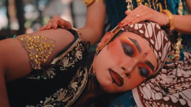 Geleneksel makyajlı bir erkeğin bir kadının kucağında dinlenmesi. Elleri, kırmızı ojeli, nazikçe başını okşuyor. Cava kültüründe sevginin, şefkatin ve şefkatin güçlü bir tasviri.