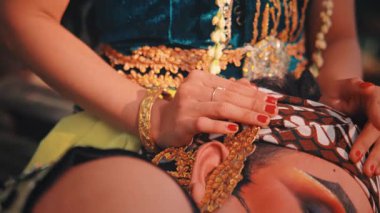 Bir kadının ellerini kırmızı oje ve altın bileziklerle gösteren aşırı yakın plan, geleneksel bir Javalı batik başlık (blangkon) takan ortağının kafasını nazikçe okşayan bir kadın.).