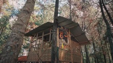 Geleneksel Endonezyalı kostümlü bir çiftin ağaç evin balkonunda romantik bir anın tadını çıkardığı düşük açılı bir çekim. Güneş etrafındaki çam ağaçlarının arkasında parlıyor ve büyülü bir atmosfer yaratıyor..