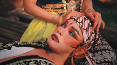 Dramatik makyajlı, başını partnerinin kucağında dinlendiren erkek bir Cava sanatçısının sanatsal yakın çekimi. Kafasına nazikçe dokunduğu ve derin duygular taşıdığı için yüzündeki ifadeye odaklanıyor..