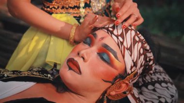 Geleneksel tiyatro makyajlı, huzur içinde dinlenen bir adamın yakın çekimi. Altın bileziklerle süslenmiş bir kadının elleri, nazikçe başını okşar, ki o da batik bir başlığa sarılır. Saf şefkat sahnesi.