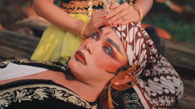 Yüksek açılı bir bakış açısı, geleneksel Cava makyajlı bir adamın başını dinlendirirken bir kadının nazikçe onu okşadığını gösteriyor. Bu tiyatro sahnesinde güzel kostümler, makyaj ve duygusal bakım ve konfor teması vurgulanıyor..