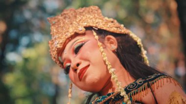 Cava dansçısı bir kadının zarif yakın plan portresi. Huzurlu ifadesi, özenle hazırlanmış makyajı ve süslü altın başlığı, Endonezya 'nın kültürel güzelliğini gözler önüne seren yumuşak odaklı bir orman geçmişine güzelce yansıtılıyor..