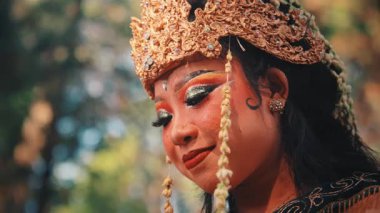 Dişi bir Cava dansçısının zarif bir yakın plan portresi. Sakin ve aşağıya bakan bir bakışı var. Çekim, Endonezya kültürünün zarafetini ve güzelliğini gözler önüne seren özenle hazırlanmış makyajını ve süslenmiş altın başlığını vurguluyor..