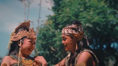 Endonezyalı mutlu bir çift geleneksel kostümler giyip, makyajlı kahkahalar atıyor ve dışarıda sohbet ediyorlar. Erkek, kadınla şakalaşarak iletişim kuruyor ve ikisi de merak içinde başını kaldırıp güzel bir kültürel an sergiliyor..