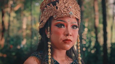 Orman ortamındaki kadın bir Cava dansçısının duygusal yakın plan portresi. Süslü, altın bir başlık takıyor ve geleneksel makyaj yapıyor. İfadesi şaşkınlık, şok ve korku dolu bir ifadeye dönüşüyor..