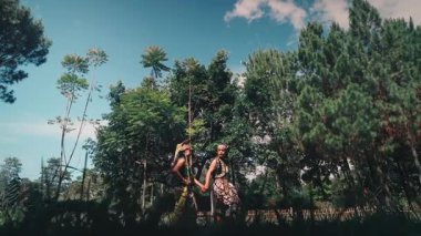 Geleneksel bir Cava çiftinin, açık bir alanda, uzun ağaçlarla birlikte beyaz bulutlu güzel mavi gökyüzüne karşı durduğu geniş açılı bir görüntü. Görkemli ve romantik bir kültürel sahne..