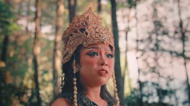 Süslü, altın başlıklı ve geleneksel kostümlü bir kadın Cava 'lının orta boy fotoğrafı. Endişeli ve duygusal bir ifadeyle bir ormanda duruyor, sanki birini arıyor ya da kaybolmuş gibi..