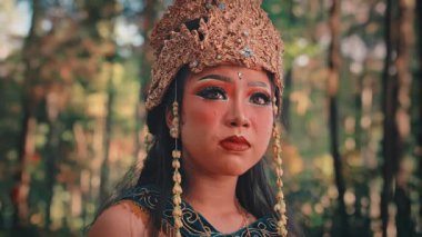 Orman ortamındaki kadın bir Cava sanatçısının duygusal yakın plan portresi. Süslü, altın bir başlık takıyor ve geleneksel makyaj yapıyor. Yüzündeki ifade endişe, korku ve endişe dolu..
