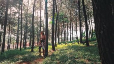 Geleneksel Endonezya okçusu gibi giyinmiş altın bir yay tutan bir adam, güneşli bir çam ormanını keşfediyor. Doğal bir yolda yalınayak yürüyor, doğa, tarih ve folklor ile bir bağ kuruyor..