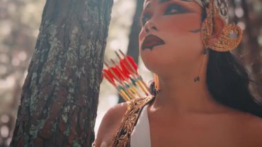 Dramatik makyajlı erkek bir Cava okçusunun yakın plan portresi, sırtında batik bir başlık ve oklar. Güneşli bir ormanda, mitolojik bir kahramanı somutlaştıran bir ağacın yanında düşünceli bir şekilde duruyor..