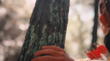 Geleneksel bir Cava okçusunun çam ağacının kabuğuna dokunuşunun yakın çekimi. Dramatik bir makyaj yapıyor ve sırtında oklar var. Gizemli, güneşli bir ormanın derinliklerine bakıyor..