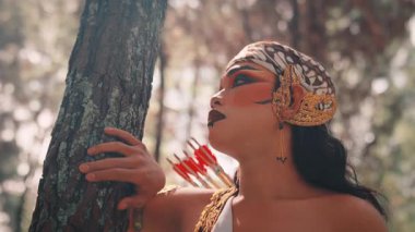 Ormandaki erkek bir Cava okçusunun sanatsal yakın çekimi. Eli ağaçta dinleniyor. Geleneksel makyaj ve batik başlık takıyor, düşünceli ve kararlı bir ifadeyle yukarı bakıyor..