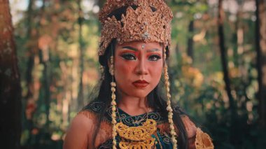 Dişi bir Cava müzisyeninin duygusal yakın çekimi. Süslü, altın bir başlık ve geleneksel bir makyajla, bir orman ortamında endişe ve üzüntü ifadesiyle etrafına bakıyor..