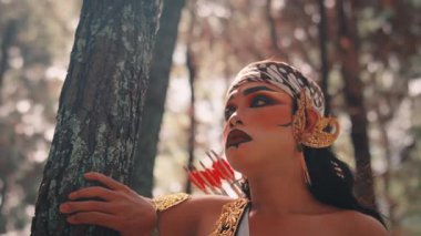 Ormandaki erkek bir Cava okçusunun sanatsal yakın çekimi. Eli ağaçta dinleniyor. Geleneksel makyaj ve batik başlık takıyor, düşünceli ve dramatik bir ifadeyle yukarı bakıyor..