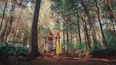 Güzel bir orman açıklığında geleneksel bir Cava giyim standında yukarı bakıp gökyüzünü gösteren bir çift. Endonezya folkloru, hikaye anlatımı ve harikalar yaratan geniş, manzaralı bir çekim..