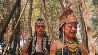 Ormanda bambu direkleri arasında dikilen geleneksel giyimli ve makyajlı bir Java çifti orta ölçekli bir fotoğraf. Etrafa dramatik ifadelerle bakınıyorlar, Endonezya folklorundan bir sahne canlandırıyorlar..