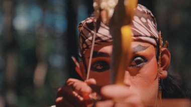 Dramatik makyajlı, ok ve yay doğrultmuş bir Cava okçusunun yoğun yakın çekimi. Onun kararlı ifadesi ve geleneksel silahının ayrıntıları üzerinde duruluyor. Folklordan bir savaşçı tasvir ediliyor..