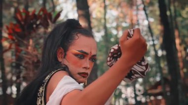 Tiyatro makyajlı, Batik mendili sıkarken üzgün bir ifadeyle yukarı bakan dramatik bir Cavalı adam. Bu duygusal performans bir ormanda geçiyor, keder ya da umutsuzluğa işaret ediyor..