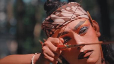 Dramatik makyajı ve batik başlığı olan odaklanmış bir Cava okçusunun yakın çekimi. Kırmızı uçlu bir oku ustalıkla fırlatır ve güneşli bir ormanda okuyla nişan alır. Efsanevi bir savaşçıyı somutlaştırır..