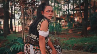 Dramatik makyajlı ve geleneksel Cava kostümlü bir adam, Endonezya 'nın zengin kültürel performansını sergilemek için ormanda bambu platformunda otururken duygusal ve güçlü bir dans sergiliyor..