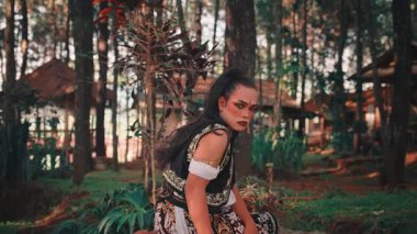 Dramatik makyajlı ve geleneksel Cava kostümlü bir adam ormandaki bir bambu platformunda oturur ve göstermeden önce yoğun bir şekilde kameraya bakar. Çekim çok güçlü ve karakterli..