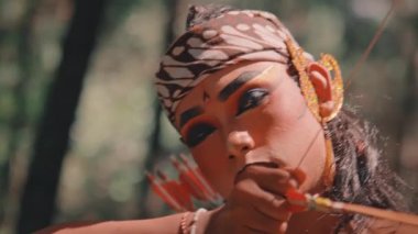 Tiyatro makyajlı, ok ve yay hedefleyen bir Cava okçusunun yoğun yakın çekimi. Onun kararlı ifadesi ve geleneksel silahının ayrıntıları üzerinde duruluyor. Folklordan bir savaşçı tasvir ediliyor..