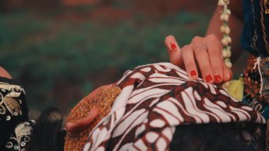 Yakın plan çekimde, bir kadının elleri kırmızı ojeyle partnerine geleneksel batik başlık (blangkon) bağlarken dikkatlice görülüyor. Samimi bir bakım, hazırlık ve kültürel gelenek sahnesi.