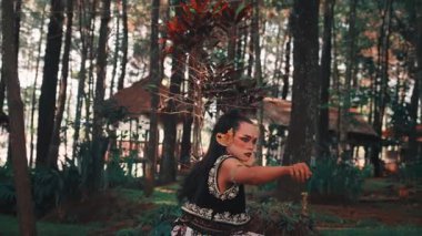 Geleneksel kostüm ve makyajlı bir erkek Cava dansçısı, çam ormanındaki bir bambu platformunda yoğun bir bakışla son bulurken güçlü ve şiddetli bir dans sahnesi sergiliyor..