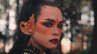 Geleneksel makyajlı ve altın küpeli bir japon erkek dansçı omzunun üzerinden yoğun bir şekilde görünüyor. Kamera yavaşça geri çekiliyor, sakin ve bulanık ormanı ortaya çıkarıyor..