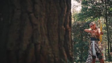 Geleneksel kıyafetli bir Cava savaşçısı, yemyeşil bir ormanda büyük bir ağaç tarafından kısmen gizlenmiş olan altın yayını hedefler. Atış, avlanma ya da savaşma konusunda bir şüphe, odaklanma ve beceri hissi yaratır..