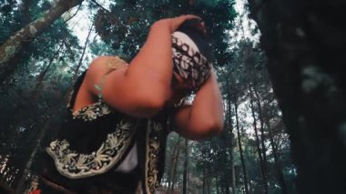 Alçak bir açıdan, süslü kostümlü bir Javalı erkek dansçı geleneksel batik başlığını ayarlar ve bir ağacın yanında anlamlı kol hareketleri yapar, sanki bir ayinmiş gibi yukarı bakar..