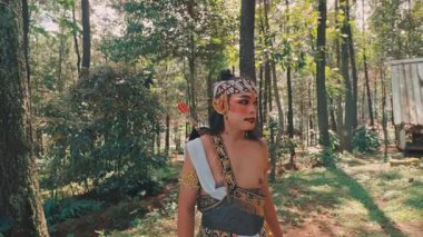 Geleneksel okçu gibi giyinmiş bir Cava erkek dansçısı güneşli bir orman açıklığında tetikte bekliyor. Etrafını dikkatli bir ifadeyle inceliyor, mitolojik bir kahramanın ruhunu somutlaştırıyor..