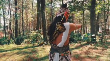 Arkadan, okçu kostümlü bir Cava dansçısı, güneş tepedeki gölgeliklerde parıldarken doğayla ruhani ve görkemli bir bağ kuran uzun ağaçlara doğru bakar..