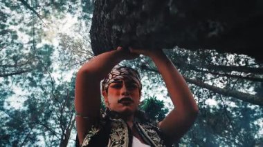 Bir ağaca tutunup orman tepe örtüsüne karşı etkileyici hareketler sergileyen dramatik makyajlı bir Cava erkek dansçısının yaratıcı düşük açılı (solucan bakışı) görüntüsü gerçeküstü ve güçlü bir görsel yaratıyor..