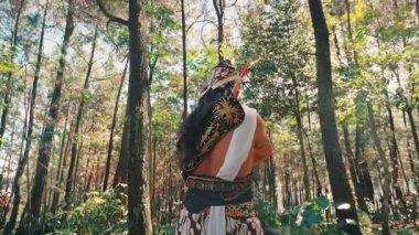 Arkadan, okçu kostümlü bir Cava dansçısı, güneş tepedeki gölgeliklerde parıldarken doğayla ruhani ve görkemli bir bağ kuran uzun ağaçlara doğru bakar..