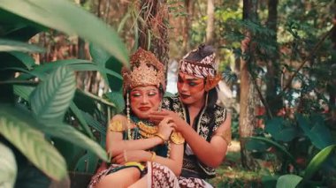 Geleneksel giyinmiş bir Java çifti arasında büyüleyici ve eğlenceli bir an. Çiçeklerin arasında oturmuş, gülüyorlar ve erkeğin kadına sevgiyle fısıldadığı gibi gülüyorlar..